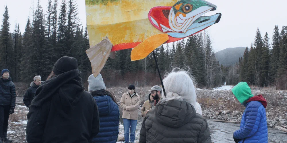 An image of people huddling in a circle in front of a river. One person is holding a multicolored cardboard fish