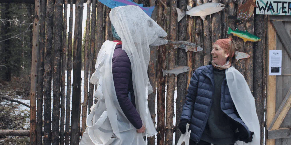 An image of two women dressed as fish standing in front of a wooden palisade
