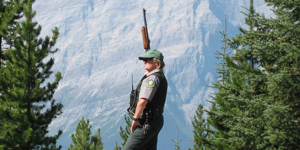 Christine Scotland stands with a rifle in front of a mountain in her uniform.