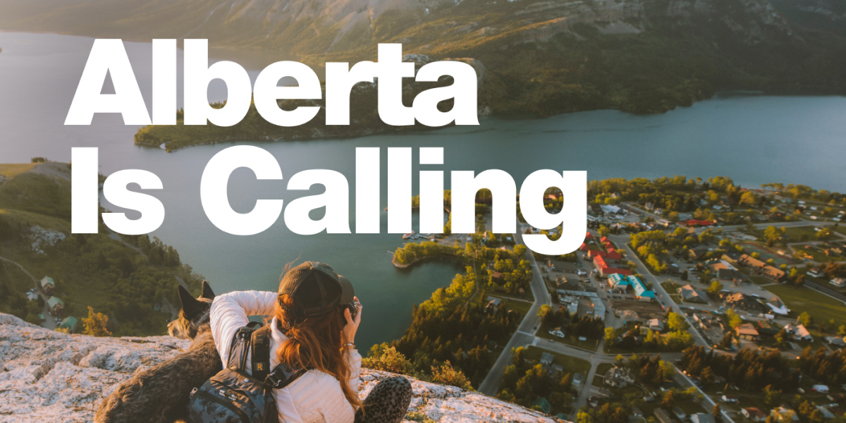 an ad for the Alberta is Calling campaign featuring a picture of a woman with her dog on top of a mountain taking a picture of Waterton, Alberta below her that is surrounded by Waterton Lake and lush greenery