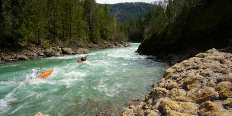 a beautiful shot of Bull River featuring flowing crystal blue water surrounded by rocks and tall trees with two kayakers traversing the water