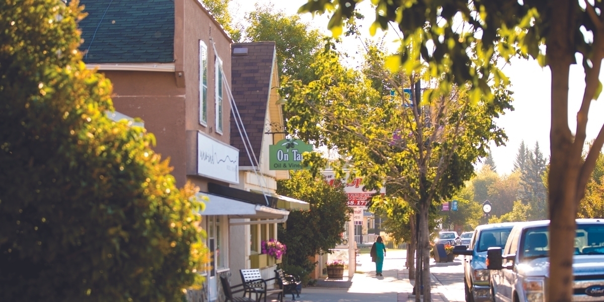 A picture of a quiet street in Downtown Okotoks showing off lush green trees and a couple of shops