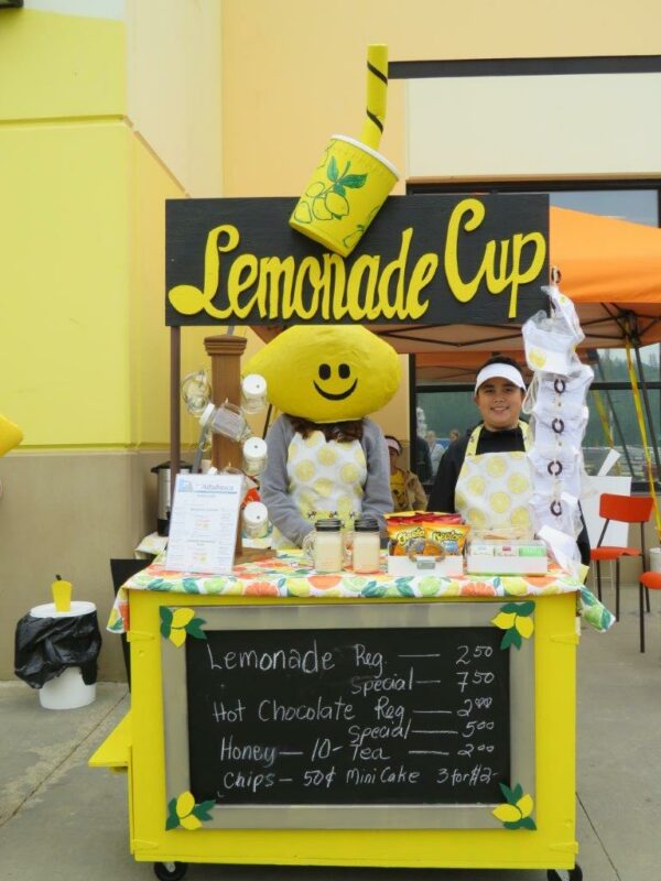 a lemonade stand from the 2019 lemonade day with a menu listing offered items and a women with a lemon costume