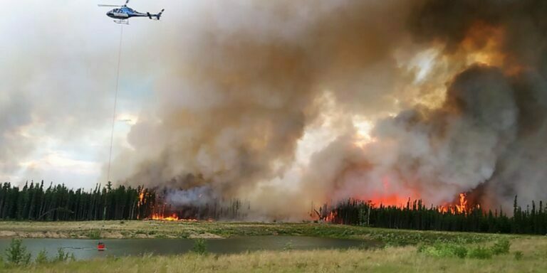 a photo of a helicopter with a bucket collecting water from a pond to douse a raging wildfire seen in the background