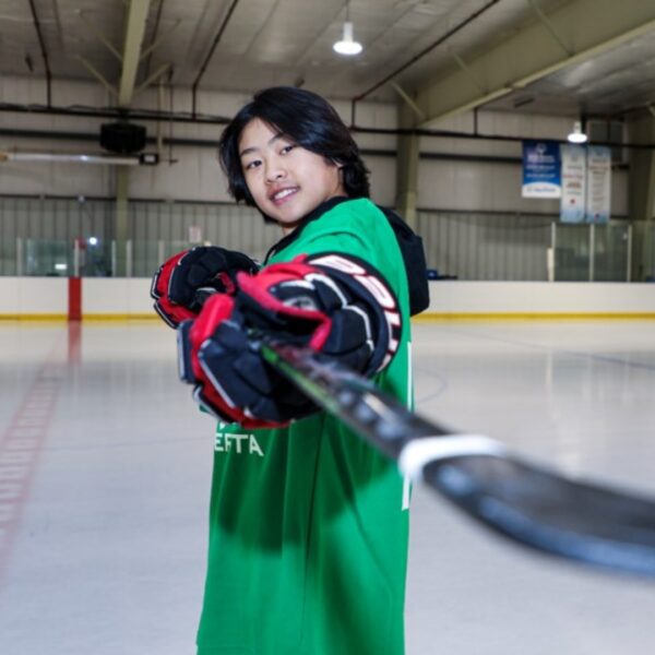 soma furue pointing a hockey stick at the camera smiling wearing a green hockey sweater