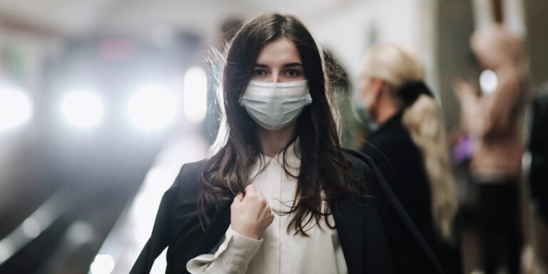 photo of a gird in a subway station wearing a medical mask with a train approaching in the background