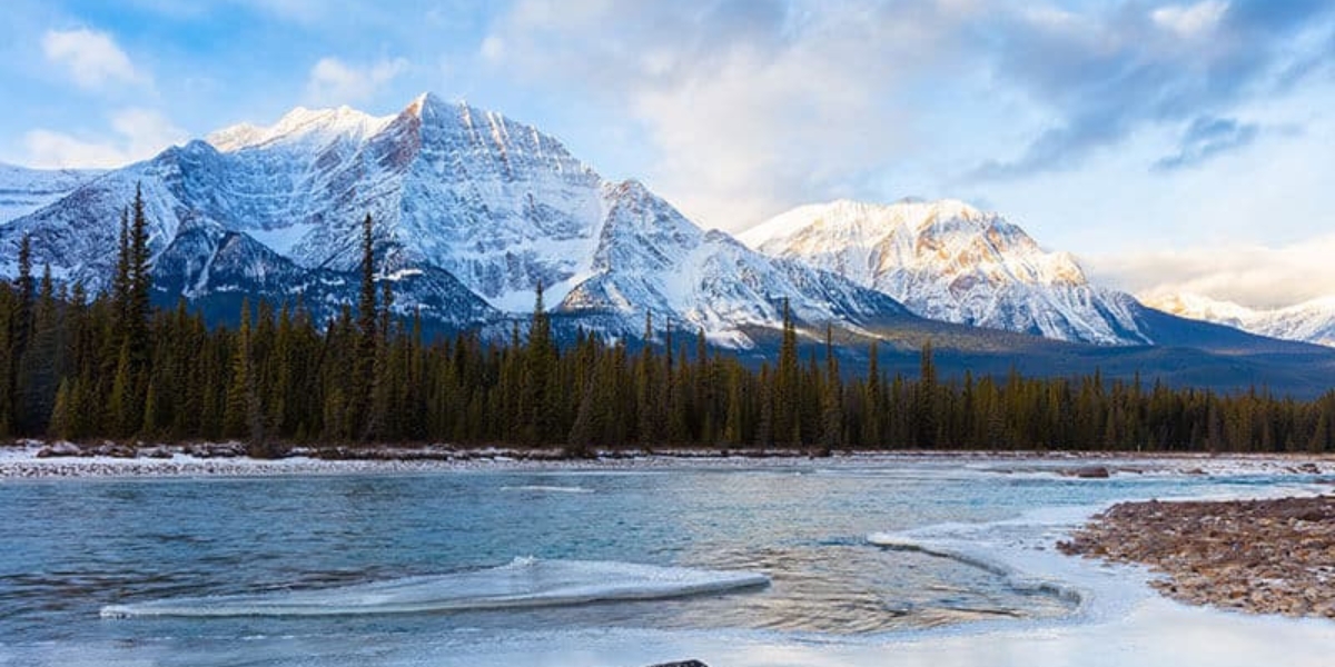 a beautiful shot of jasper national park in the winter with a river in the forefront and trees and the rockies in the distance