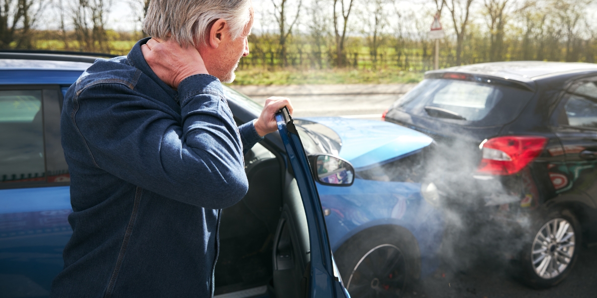 a man in a jean jacket holding his neck holding on to the door of his car which rear ended another vehicle with a country landscape in the distance