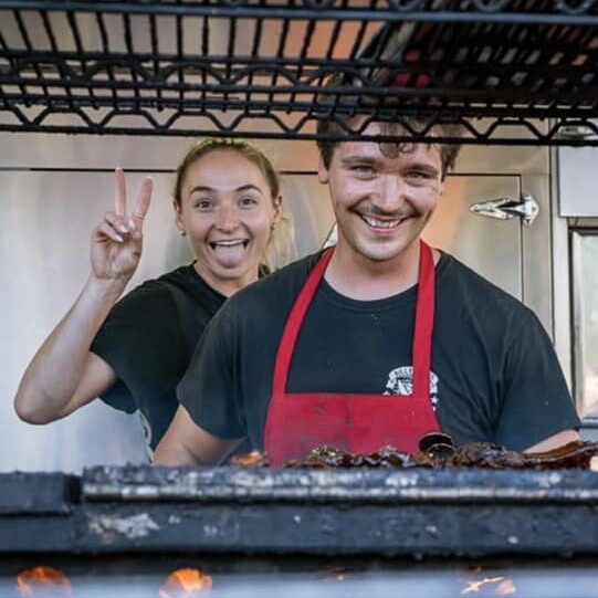 a young man behind a grill cooking ribs with a girl behind him holding up the peace sign with her fingers