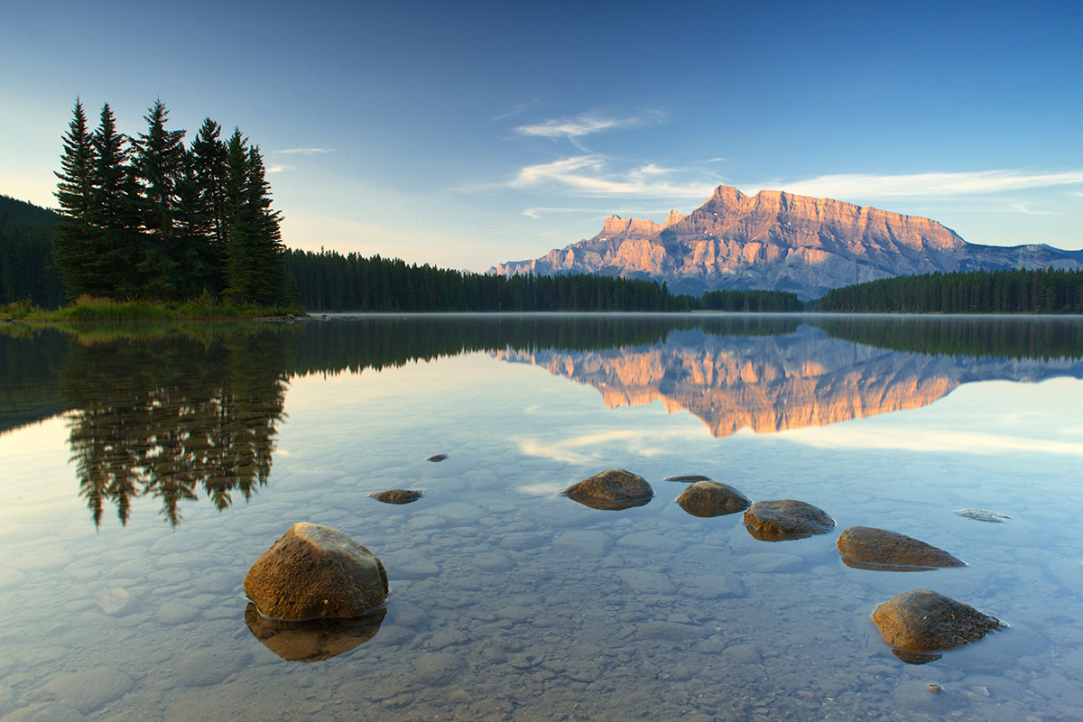 Photograph of a mountain lake with rocks in the foreground and a mountain at sunrise, all reflected in calm waters.