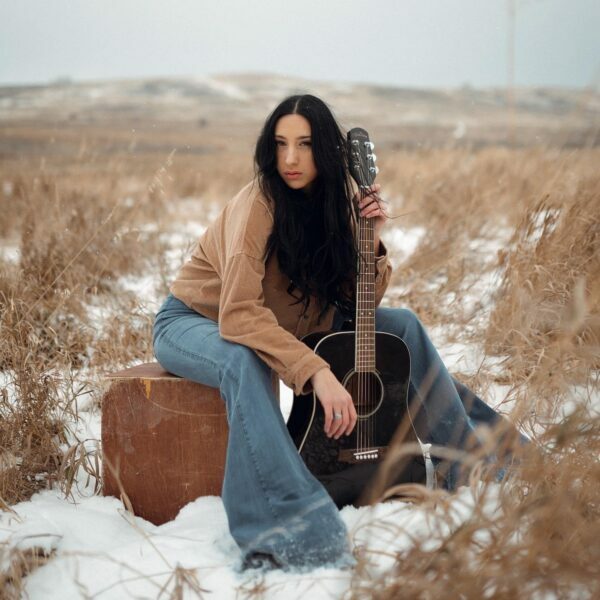 a photo of ashley ghostkeeper sitting on a wooden box holding her black guitar in an open brown field