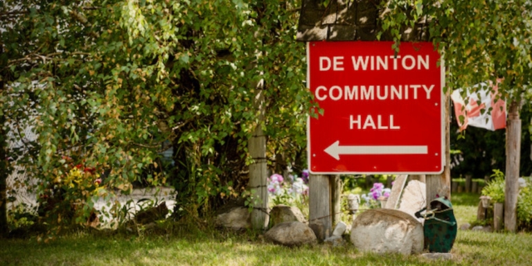 a red sign that reads de winton community hall surrounded by lush greenery