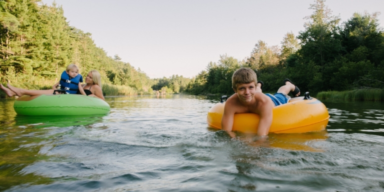 a boy on a yellow tube and a woman holding a child wearing a blue life jacket in a green tube on the water surrounded by trees