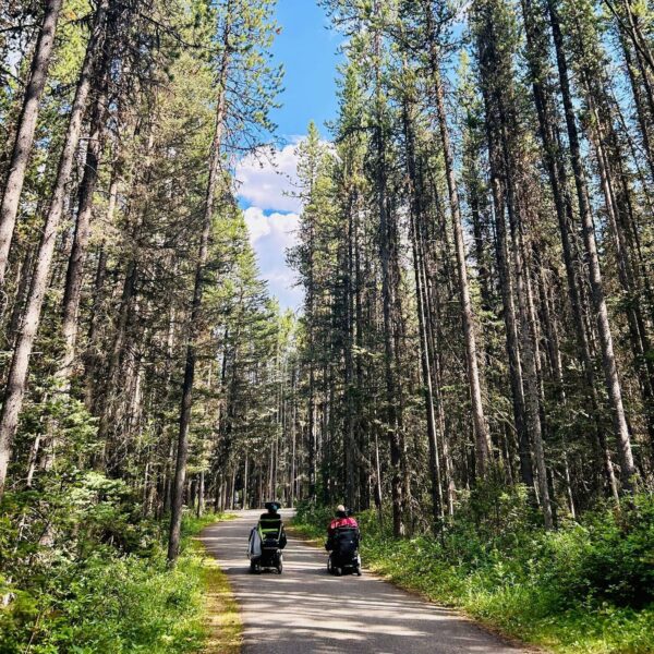 Two people in 2 wheelchairs navigating a paved pathway in the forest in Kananaskis