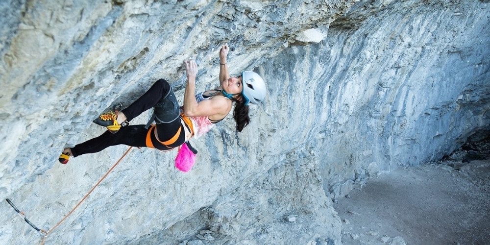 sport climber becca frangos scaling a steel rock wall wearing a pink top and leggings equipped with safety gear looking determined