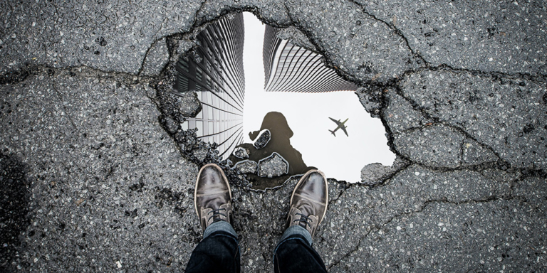 Photo of a pothole filled with water with a person's shoes and legs standing over the pothole. There is a reflection of city skyscapers and an airplane in the pothole.