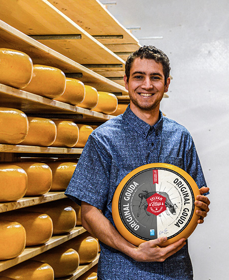 photo of man holding a ring of gouda with shelves of goda chesse in the background