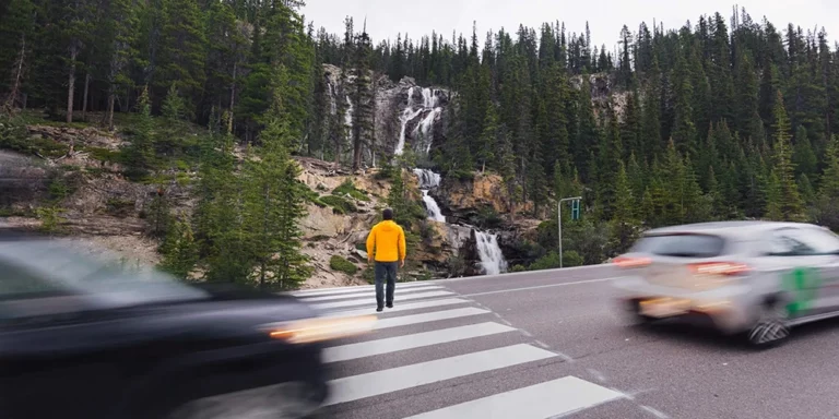 picture of a man at a crosswalk in the mountains walking toward a waterfall alongside the road with cars on the highway