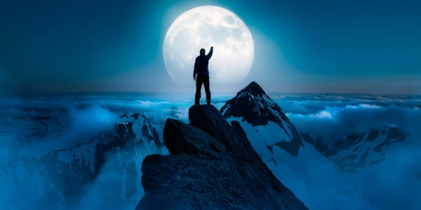 Man standing on a mountain oeak with hand up silhoutted by the full moon