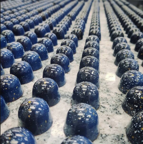 Many glossy chocolate bonbons. They are a darker shade of indigo in colour and have several while dots on them. 